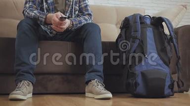 一个坐在沙发上的男人按下<strong>电视</strong>遥控器的按钮，旁边是一个大<strong>旅游</strong>背包